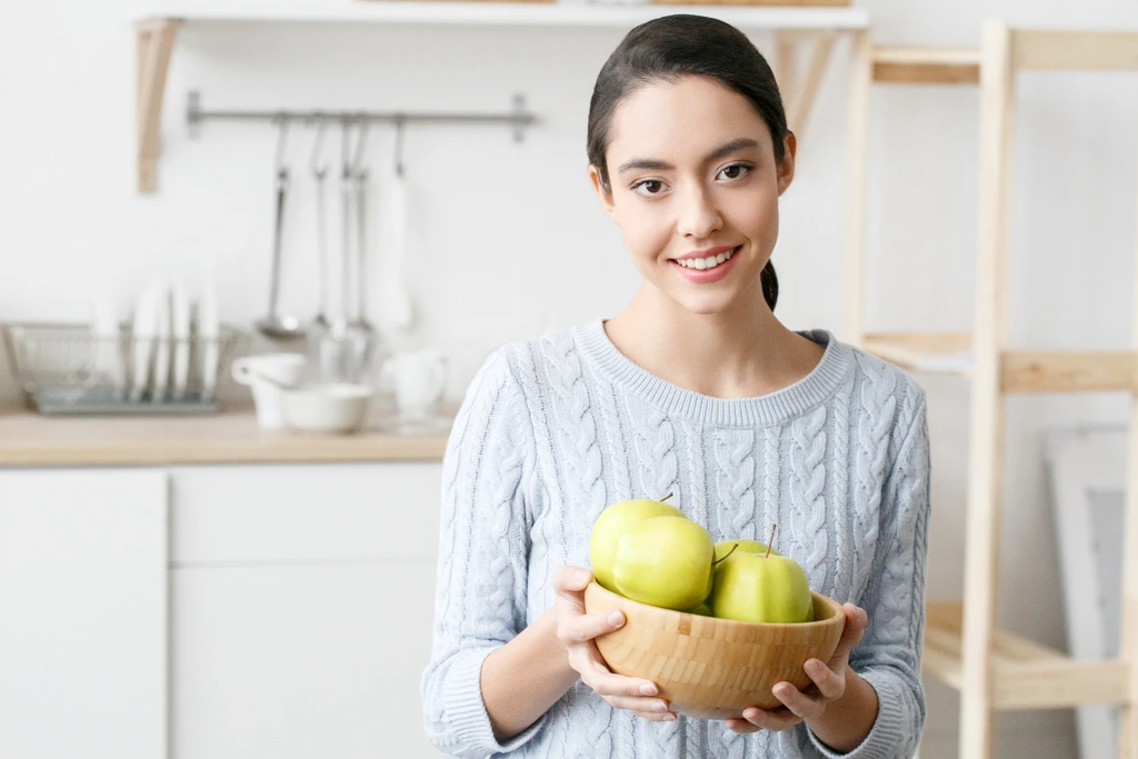 Ava Sinclair, author of Misty Plate, smiling in her bright white kitchen while holding a wooden bowl filled with green apples.