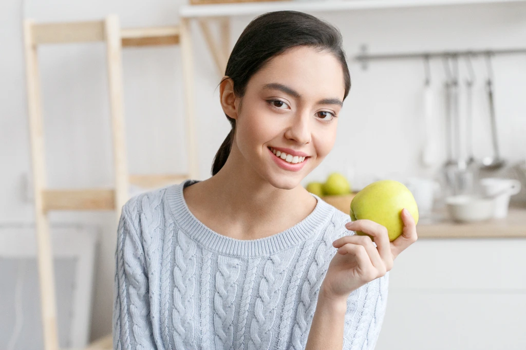 A friendly portrait of Ava Sinclair from the recipe blog Misty Plate, smiling as she holds up a single green apple in her kitchen.