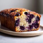 Close-up of a slice of moist blueberry banana bread showing the golden crust and purple blueberries baked into the crumb.