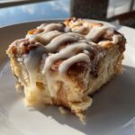 A close-up, sunlit photo of a single serving of fluffy Cinnamon Roll Casserole topped with thick, dripping white icing.