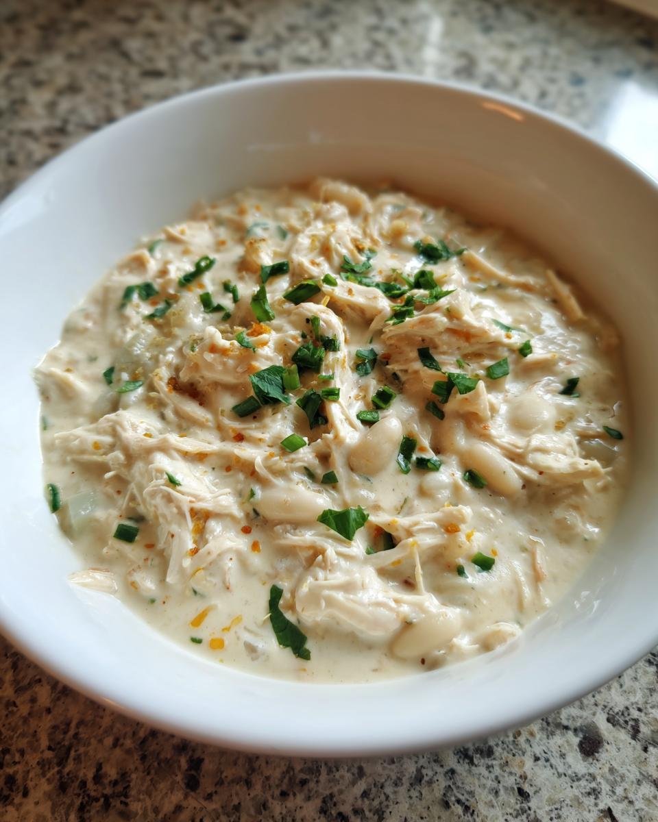 A close-up of a white bowl filled with creamy crockpot white chicken chili, featuring shredded chicken, white beans, and parsley garnish.