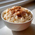 A close-up of creamy rice pudding served in a white bowl, generously sprinkled with ground cinnamon.