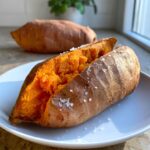 A fluffy baked sweet potato, split open and sprinkled with coarse sea salt, sitting on a white plate.