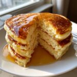 A thick stack of fluffy pancakes with a piece cut out, showing the airy interior and drizzled with golden syrup.
