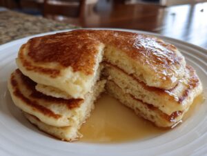 Close-up of a stack of three fluffy pancakes with a slice cut out, showing the airy interior and drizzled with golden syrup.