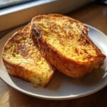Two thick slices of golden brown easy french toast resting on a speckled white plate, illuminated by sunlight.