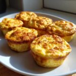 Six golden brown, fluffy high protein egg bites arranged on a white plate, catching bright sunlight.