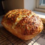 A freshly baked loaf of Italian herb and cheese bread with a golden, cheesy crust and visible herbs, resting on a wire rack.