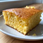 Close-up of a square slice of moist buttermilk cornbread with a golden-brown, shiny top, served on a white plate.