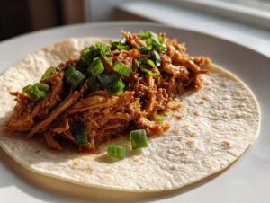 A soft tortilla topped with saucy shredded chicken filling and chopped green onions, ready to be made into chicken tacos.