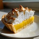 A cross-section slice of lemon meringue pie showing bright yellow curd and tall, toasted meringue peaks.
