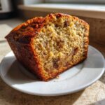 A close-up shot of a moist slice of sugar free banana bread on a white plate, bathed in sunlight.