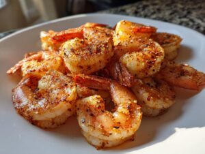 Close-up of cooked, seasoned air fryer frozen shrimp piled on a white plate, showing a crispy, reddish-brown spice coating.