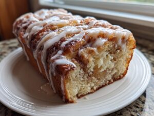 A whole loaf of amazing apple fritter bread drizzled generously with white icing, sitting on a white plate.