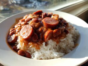 A close-up of a white bowl filled with white rice topped generously with savory beans and rice stew and sliced smoked sausage.
