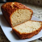 A golden brown loaf of homemade beer bread, with one thick slice cut and resting next to the main loaf on a white plate.