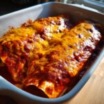 Close-up of baked cheese enchiladas covered in melted orange cheese and rich red sauce in a white baking dish.