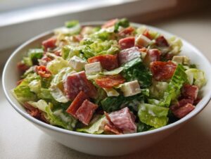 A close-up of a hearty grinder salad featuring chopped lettuce, cubes of meat, cheese, and creamy dressing in a white bowl.