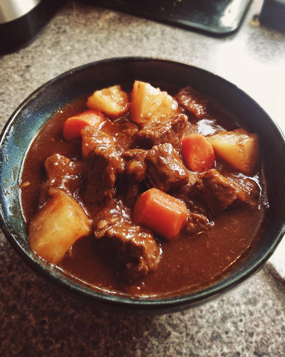 A close-up of a dark bowl filled with rich, dark brown pork stew, featuring chunks of meat, potatoes, and carrots.