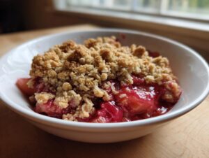 A close-up of a serving of amazing rhubarb crisp in a white bowl, showing the bright red fruit filling and golden brown crumble topping.