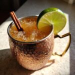 Close-up of an Apple Cider Moscow Mule cocktail in a hammered copper mug, garnished with a lime wedge and cinnamon stick.