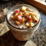Close-up of apple cinnamon overnight oats in a glass jar topped with diced red apples and cinnamon.