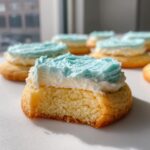 Close-up of a soft, golden sugar cookie with a bite taken out, topped with white and bright blue frosting.