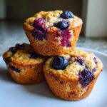 Three golden-brown blueberry cottage cheese muffins stacked on a white plate, highlighting the burst blueberries.