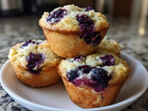 A stack of three golden-brown blueberry cottage cheese muffins topped with burst blueberries, resting on a white plate.