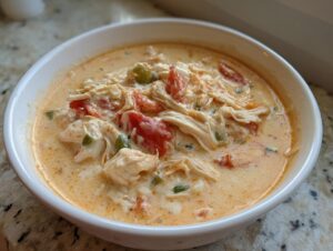 Close-up of a white bowl filled with creamy, orange-hued cream cheese chicken chili, featuring shredded chicken, tomatoes, and green peppers.