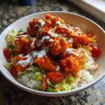 A close-up of a delicious buffalo chicken bowl featuring crispy buffalo-coated chicken over rice, lettuce, tomatoes, and drizzled with ranch dressing.