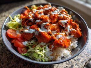 A close-up of a buffalo chicken bowl featuring saucy chicken pieces over rice, lettuce, tomatoes, and drizzled with ranch dressing.