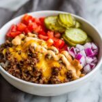 A close-up view of a delicious cheeseburger bowl featuring seasoned ground beef, melted cheddar cheese, special sauce, diced tomatoes, pickles, and red onions.