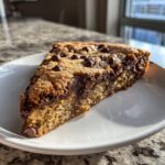 A close-up slice of a perfectly baked, chewy chocolate chip cookie cake on a white plate.
