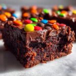 A close-up, moist slice of cosmic brownies topped with rich chocolate frosting and colorful candy-coated chocolates.