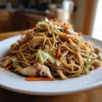A generous serving of homemade chicken chow mein recipe piled high on a white plate, featuring noodles, chicken pieces, and stir-fried vegetables.