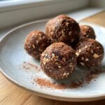 Five dark chocolate protein balls, visible with oats, dusted with cocoa powder, resting on a light plate.