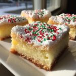 Close-up of a slice of Christmas gooey butter cookies on a white plate, topped with powdered sugar and red and green sprinkles.