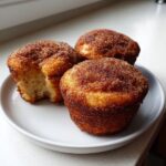 Three golden brown cinnamon sugar french toast muffins served on a white plate, one muffin is broken open showing the soft interior.