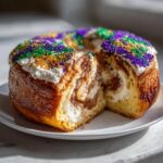 A cross-section view of a delicious king cake showing cinnamon swirl filling and topped with white icing and Mardi Gras sprinkles.