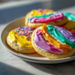 Close-up of sugar cookies topped with vibrant, rainbow-striped sugar cookie icing and sprinkles on a white plate.