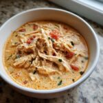 Close-up of a white bowl filled with creamy, orange-hued cream cheese chicken chili topped with shredded chicken and diced tomatoes.