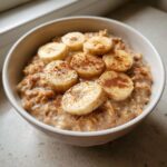 A close-up of creamy banana oatmeal in a white bowl, topped with fresh banana slices and a sprinkle of cinnamon.