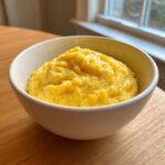 A close-up of a white bowl filled with bright yellow, creamy cheese grits sitting on a wooden table near a window.