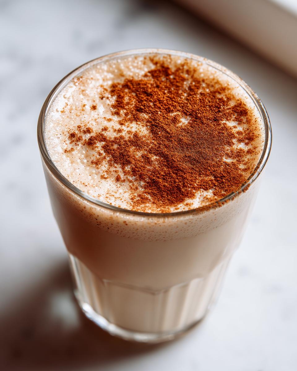 Close-up of a glass filled with creamy coquito, topped generously with ground cinnamon.