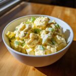 A white bowl filled with creamy, chunky egg salad recipe garnished with chopped green onions.