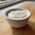 A small white ramekin filled with creamy horseradish sauce, topped with cracked black pepper.