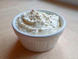 A white ramekin filled with thick, creamy horseradish sauce, topped with cracked pepper and herbs.