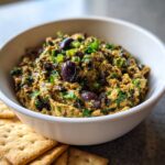 A close-up of a bowl filled with creamy olive dip, garnished with whole Kalamata olives and fresh green herbs.