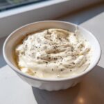 A close-up of rich, creamy olive garden alfredo sauce topped with cracked black pepper in a white bowl.